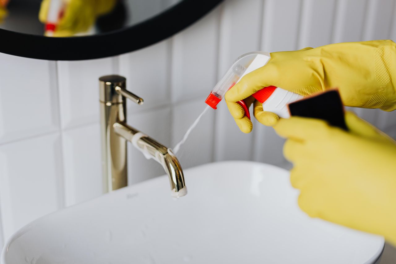 Vue d’une femme de ménage anonyme en gants en caoutchouc jaunes, pulvérisant un nettoyant sur un robinet chromé dans une salle de bain
